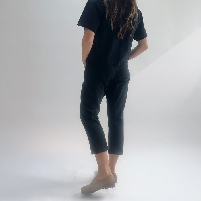 Back view of model wearing black cropped Sunday pants with hands in pockets, styled with a black short-sleeve tee and tan loafers.