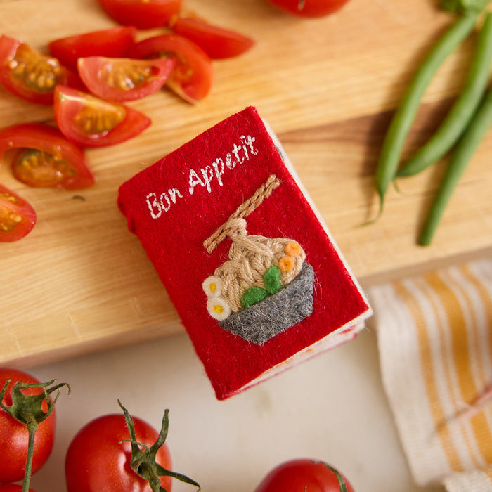 Miniature felt ornament shaped like a cookbook, hand-felted and embroidered with cooking-themed details including salt, pepper, vegetables, and a stovetop. Crafted from merino-blend wool with soft pages and a hanging string, this ornament measures approximately 2.5 inches tall. A cozy tribute to home cooking and culinary tradition.