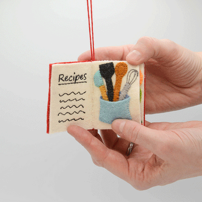Miniature felt ornament shaped like a cookbook, hand-felted and embroidered with cooking-themed details including salt, pepper, vegetables, and a stovetop. Crafted from merino-blend wool with soft pages and a hanging string, this ornament measures approximately 2.5 inches tall. A cozy tribute to home cooking and culinary tradition. 360 degree of book pages.