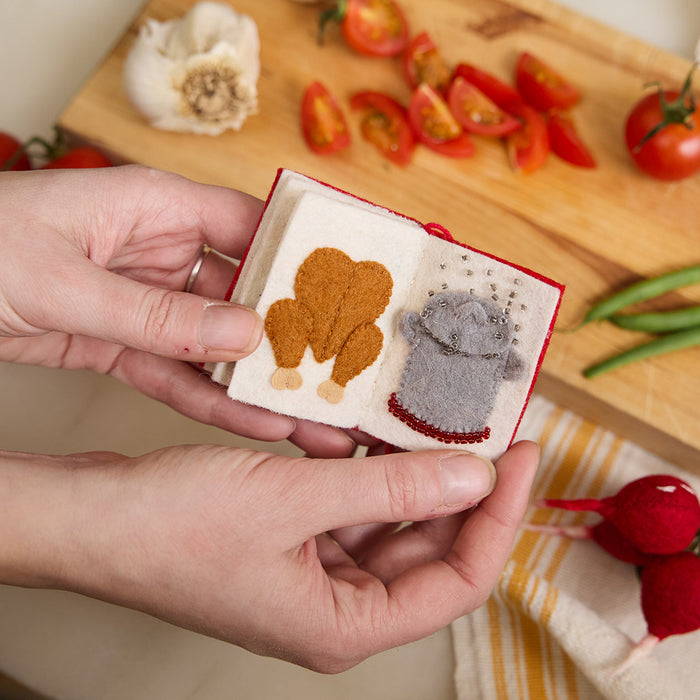 Miniature felt ornament shaped like a cookbook, hand-felted and embroidered with cooking-themed details including salt, pepper, vegetables, and a stovetop. Crafted from merino-blend wool with soft pages and a hanging string, this ornament measures approximately 2.5 inches tall. A cozy tribute to home cooking and culinary tradition. Open to page with a chicken and a boiling pot.