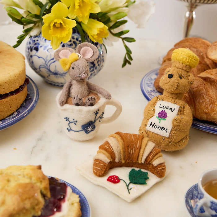 Hand-felted wool ornament of a small gray mouse with a bow, sitting inside a floral teacup with embroidered details. Crafted from merino-blend wool, this whimsical piece evokes tea time and childhood storytelling. Featured next to the honey bear bottle and croissant ornaments. Includes a hanging string and measures approximately 4.5 inches tall. A sweet and cozy decoration for tea lovers and mouse enthusiasts alike.