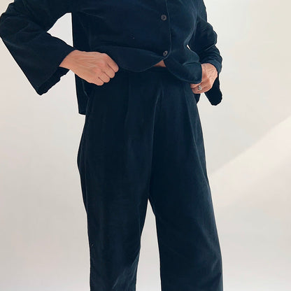 Close-up front view of the Bryn Walker Corduroy Pleated Pants in Black, showing front pleat detail, flat waistband, and black pinwheel corduroy texture, styled with matching Standing Collar Crop Jacket