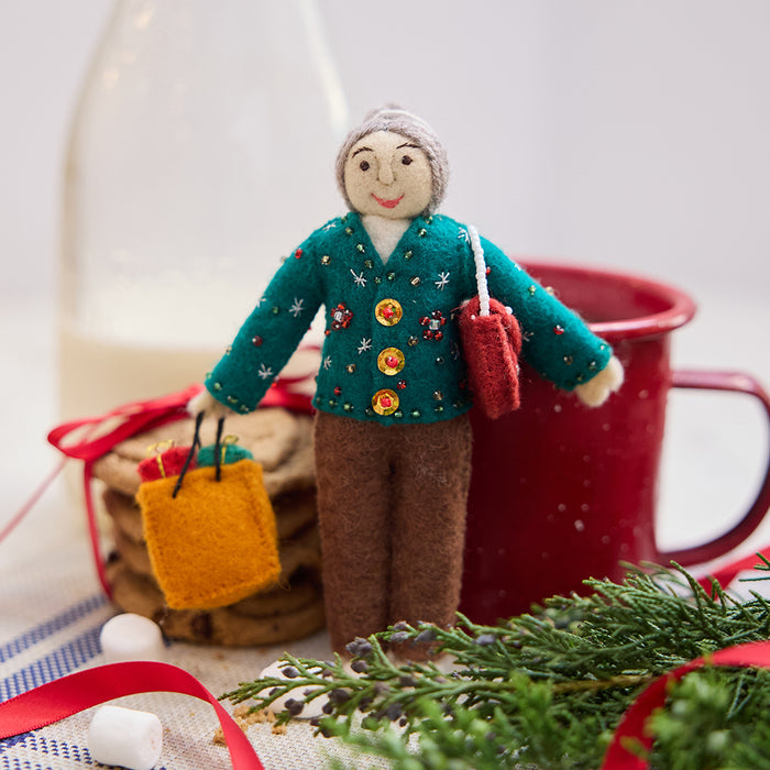 Handmade felt Grandma ornament from Craftspring, wearing a cozy beaded and embroidered holiday sweater, holding a designer purse and a shopping bag full of presents—displayed with milk, cookies, and evergreen branches, evoking warm holiday memories and timeless festive cheer, ethically crafted by artisans using sustainable wool.