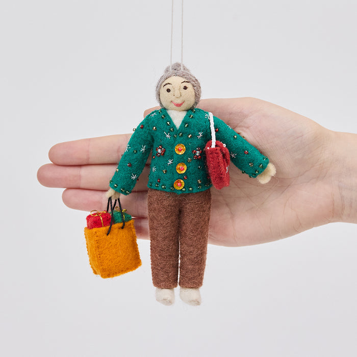 Handmade felt Grandma ornament from Craftspring held in front of a hand to show its size—featuring intricate beaded and embroidered details on a holiday sweater, a designer purse, and a shopping bag full of presents, ethically crafted by artisans using sustainable wool.