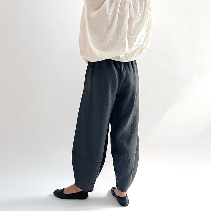 Back view of model wearing the Bryn Walker Heavy Linen Oliver Pant in Moonlight, showing elastic waistband, full barrel-leg drape, and tapered ankle in deep moonlight charcoal heavy linen, styled with cream oversized top and black ballet flats