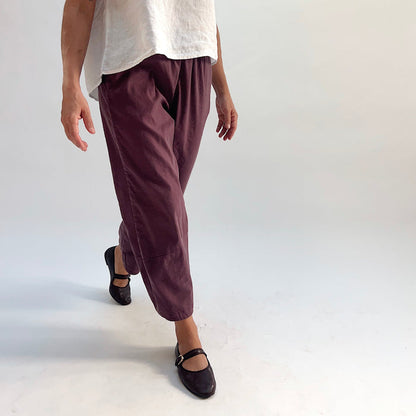 Model walking in russet linen-tencel lantern pants with curved seam legs and elastic waistband, paired with a white top and dark Mary Jane flats, side-angle view