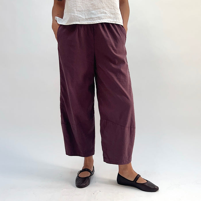 Full front view of russet linen-tencel lantern pants with wide barrel-leg silhouette, elastic waistband, side pockets, and curved hem, styled with a white linen top and dark Mary Jane flats