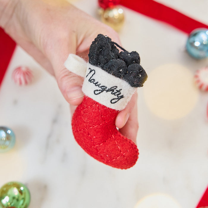 Craftspring’s Naughty Stocking with Coal Ornament — felt red stocking filled with felt coal and beads, embroidered “naughty” — held in hand over holiday baubles, capturing whimsical charm, festive warmth, and heirloom-quality Christmas gifting, ethically crafted by artisans using sustainable wool.