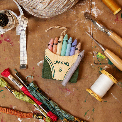 Hand-felted pastel crayon box ornament with stitched detailing, celebrating creativity and handmade craftsmanship. Perfect for teachers and young artists.