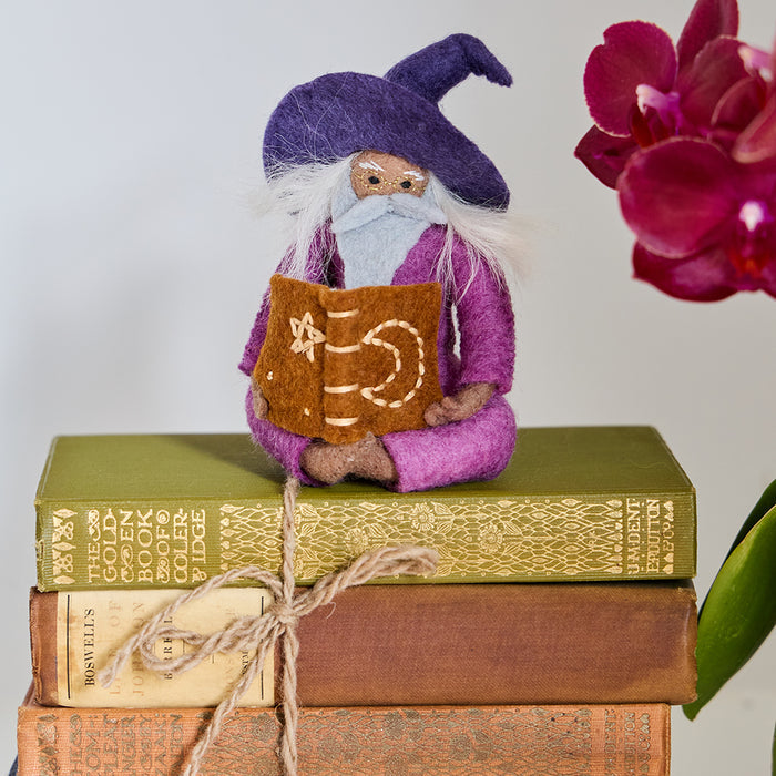 Hand-felted wool ornament of a grey-bearded wizard wearing a pointed hat, holding an open spell-book beneath swirling stardust. Featuring a warm-toned complexion, white hair, and flowing robes, this whimsical figure is crafted from natural merino-blend wool. Includes a hanging string and measures approximately 5 inches tall.