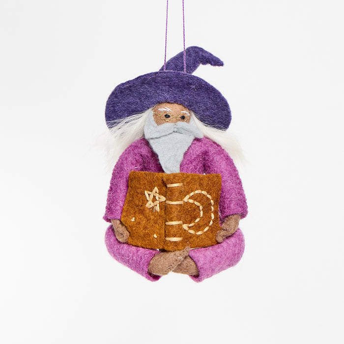 Hand-felted wool ornament of a grey-bearded wizard wearing a pointed hat, holding an open spell-book beneath swirling stardust. Featuring a warm-toned complexion, white hair, and flowing robes, this whimsical figure is crafted from natural merino-blend wool. Includes a hanging string and measures approximately 5 inches tall.