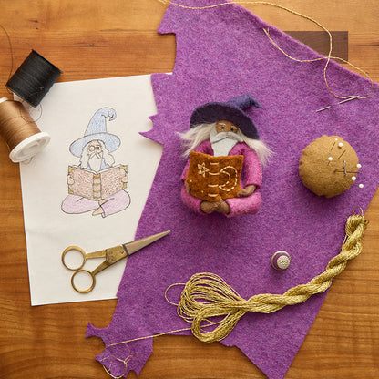 Behind the scenes look at the making of a handmade and-felted wool ornament of a grey-bearded wizard wearing a pointed hat, holding an open spell-book beneath swirling stardust. Featuring a warm-toned complexion, white hair, and flowing robes, this whimsical figure is crafted from natural merino-blend wool. Includes a hanging string and measures approximately 5 inches tall.