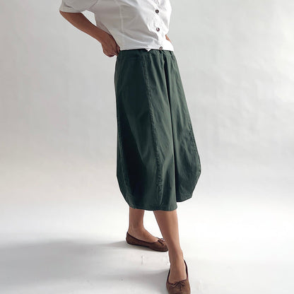 Three-quarter front view of model wearing Eleven Stitch konbu rib yoke skirt with hand on hip, showing elastic knit waistband, curved seam paneling, sculptural bubble hem, below-the-knee length, and deep forest green color, styled with white chalk camp shirt and brown ballet flats
