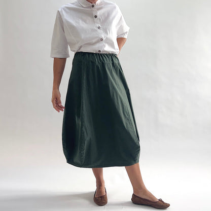 Full front view of model wearing Eleven Stitch konbu rib yoke skirt with arm at side, showing elastic knit waistband, curved seam paneling, sculptural bubble hem, below-the-knee length, and deep forest green color, styled with white chalk camp shirt and brown ballet flats