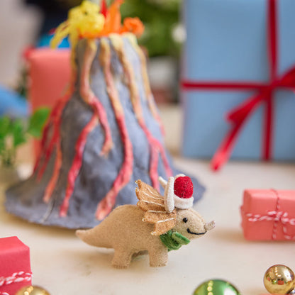 Handmade felt Santa Triceratops ornament from Craftspring, displayed in front of a volcano school project with wrapped presents nearby—perfect for dino-loving kids and joyful holiday gifting, featuring festive holiday charm with embroidered and beaded details, ethically crafted using sustainable wool.