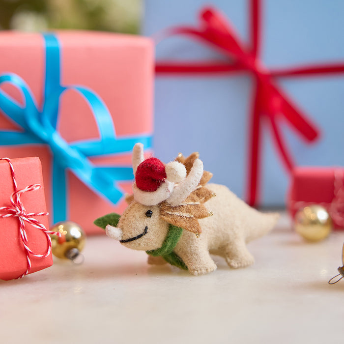 Handmade felt Santa Triceratops ornament from Craftspring, displayed in front of festive wrapped presents—perfect for dino-loving kids and joyful holiday gifting, featuring a dinosaur wearing a red Santa hat and green scarf with embroidered and beaded details, ethically crafted using sustainable wool.