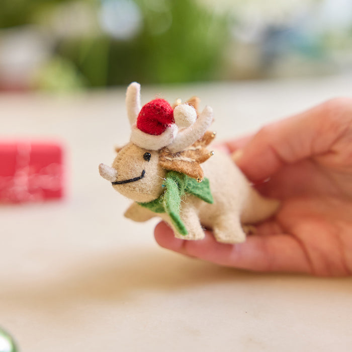 Handmade felt Santa Triceratops ornament from Craftspring held above marble with a mini wrapped present in the background—perfect for dino-loving kids and joyful holiday gifting, featuring embroidered and beaded details and a cozy red Santa hat and green scarf, ethically crafted using sustainable wool.