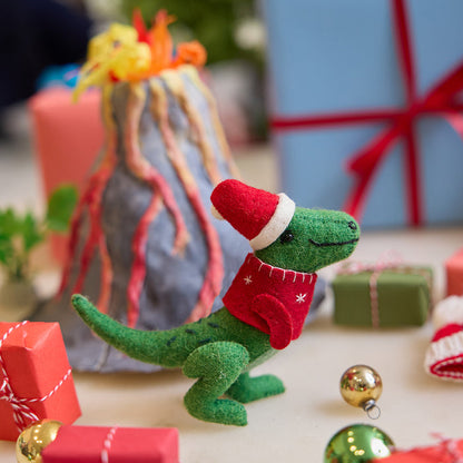 Handmade felt Santasaurus T-Rex ornament from Craftspring, displayed in front of a volcano school project with wrapped presents nearby—featuring a red Santa hat, red sweater, and embroidered details, perfect for dino-loving kids and festive holiday gifting, ethically crafted using sustainable wool.