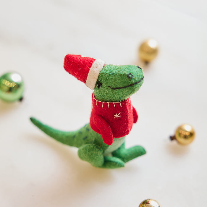 Handmade felt Santasaurus T-Rex ornament from Craftspring, displayed on marble with festive bobble ornaments—featuring a red Santa hat, red sweater, and embroidered details, perfect for dino-loving kids and cheerful holiday gifting, ethically crafted using sustainable wool.