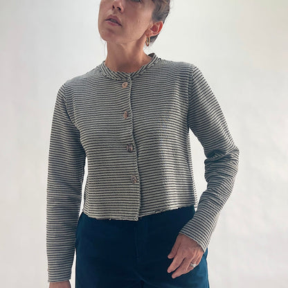 Front view of model wearing a squirrel and black fine horizontal stripe cropped cardigan with asymmetrical button closure, band neckline, and long sleeves, hand on hip, showing the crinkle-texture fabric and cropped length, styled with dark teal wide-leg pants against a white background.