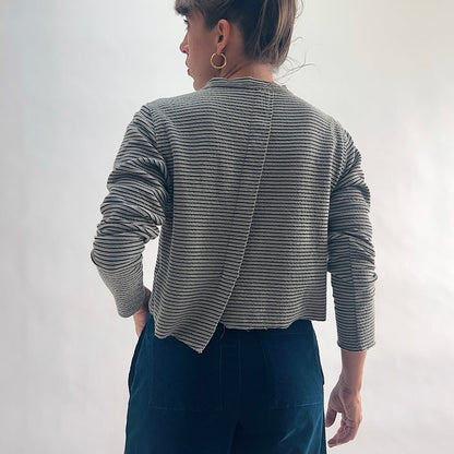 Back view of model wearing a squirrel and black fine horizontal stripe cropped cardigan with long sleeves and asymmetrical hem, hand in pocket, showing the crinkle-texture fabric and center back seam, styled with dark teal wide-leg pants against a white background.
