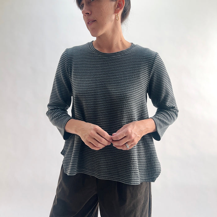 Front view of model wearing an iron charcoal and black tonal textured stripe crewneck top with 3/4 sleeves, hands clasped at center, showing the crew neckline, crinkle-texture fabric, and curved hem, styled with dark olive wide-leg pants against a white background.