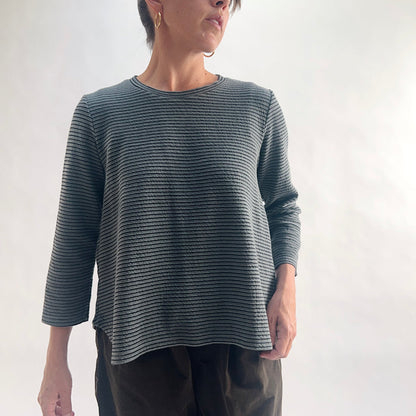 Close front view of model wearing an iron charcoal and black tonal textured stripe crewneck top with 3/4 sleeves, arm at side, showing the crew neckline, crinkle-texture fabric, and relaxed A-line silhouette, styled with dark olive wide-leg pants.