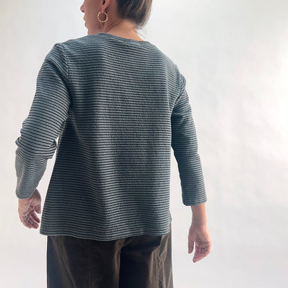 Back view of model wearing an iron charcoal and black tonal textured stripe crewneck top with 3/4 sleeves, showing the curved hem, side vents, and crinkle-texture fabric, styled with dark olive wide-leg pants against a white background.