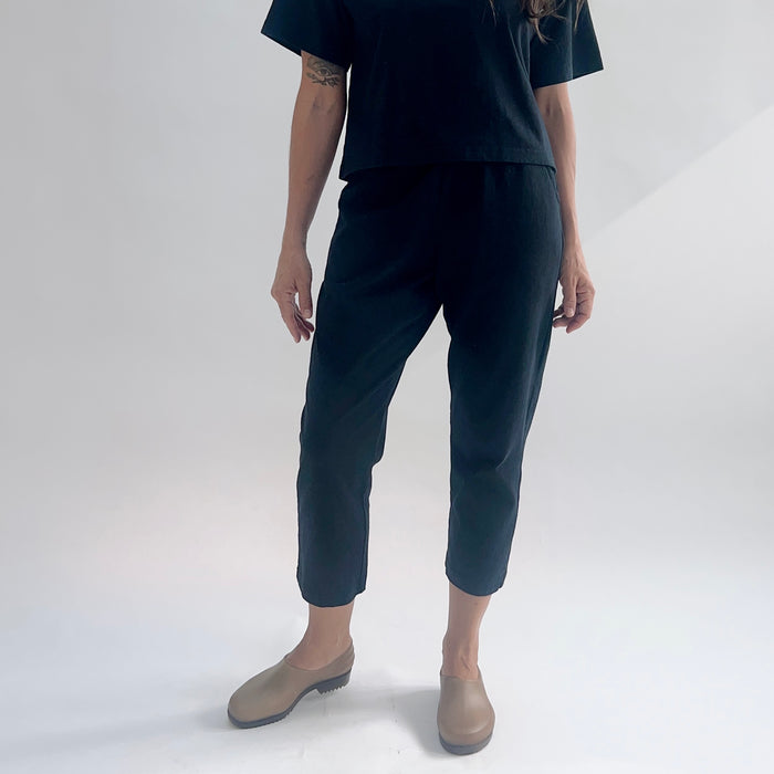 Close front view of model wearing black cropped Sunday pants with a tapered leg, styled with a black boxy tee and tan clogs.