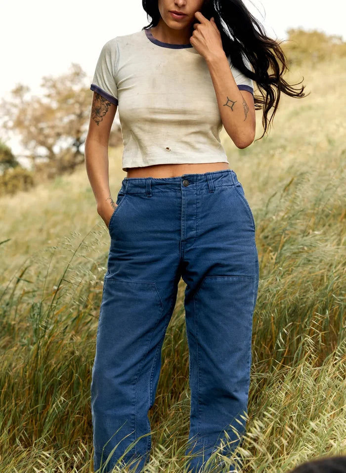 Model wearing blue high-rise Clarke Canvas Workpants with a cream ringer tee, standing in an open grassy field.